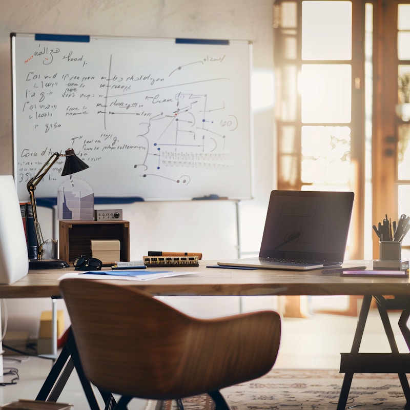 Photo: A cozy and modern home office setup featuring a wooden desk, a laptop, and a whiteboard covered with notes. Sunlight streams through the large windows, creating a warm and productive environment.