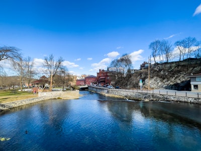 River in Park Under Blue Sky - A river with a stone wall and a building on the side