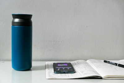 Working at Desk with Sketchbook and Coffee While Listening to a Podcast - A phone and a blue coffee tumbler on a book