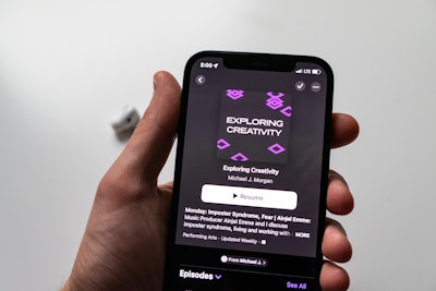 Hand Holding Phone with Podcast App - A hand holding a smartphone with a podcast on the screen and earbuds blurred in the background
