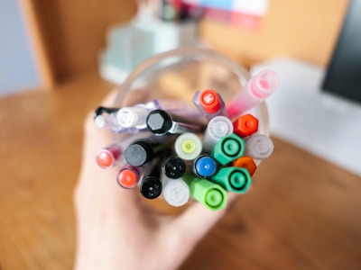 Pens and Pencils on Desk - A focused hand holding a cup of pens, markers, highlighters, and pencils over a wooden desk workspace. 