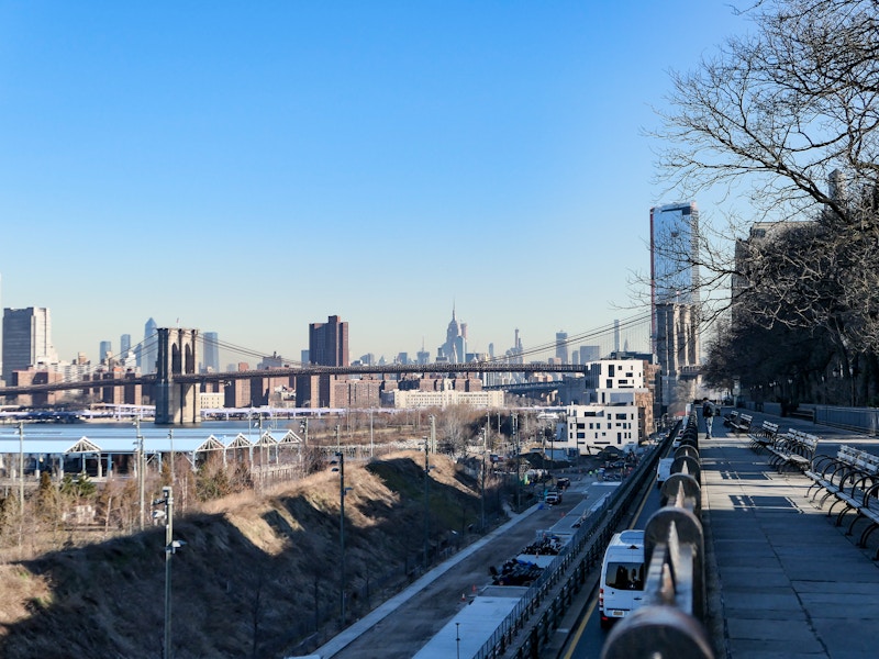 Photo: A promenade over a road with a bridge and a city in the background under blue skies 
