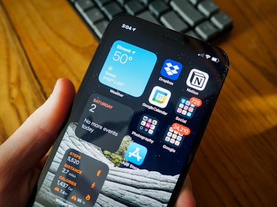 Hand Holding iPhone - A hand holding a smartphone over a blurred keyboard and wooden desk in the background 