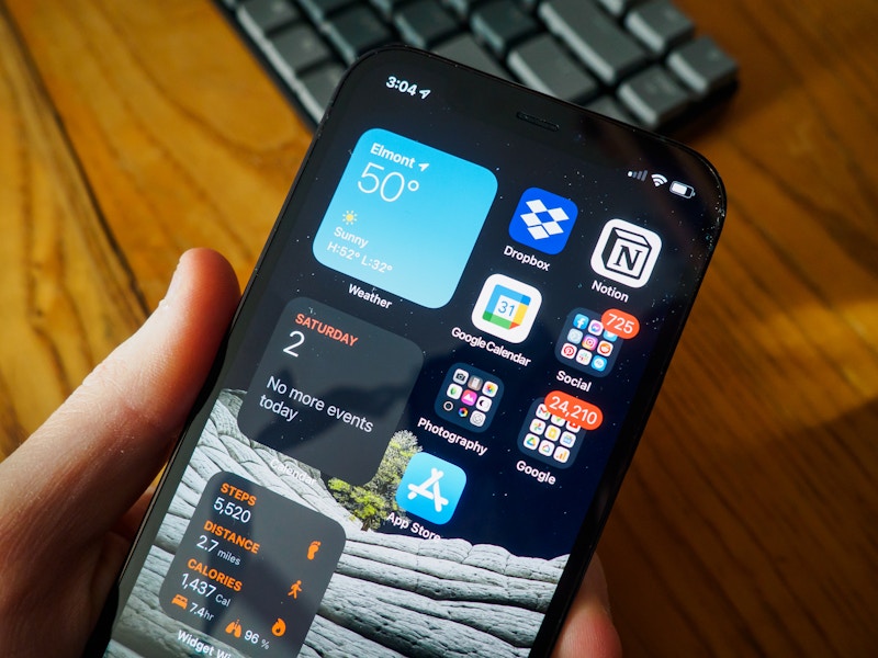 Photo: A hand holding a smartphone over a blurred keyboard and wooden desk in the background 