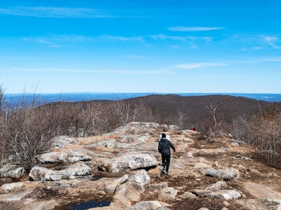People Hiking on Mountain Under Blue Sky - A person walking on a rocky mountain hill in front of a blue sky and more hills