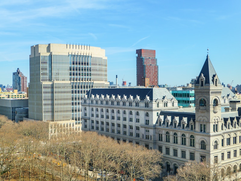 Photo: A group of buildings in a city with trees in front under blue skies 