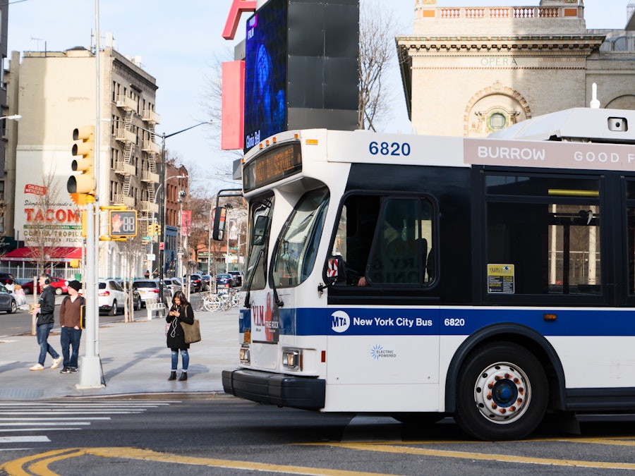 Photo: A white and blue bus on a city street with people and buildings 