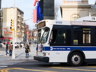 New York City Sidewalk and Bus - A white and blue bus on a city street with people and buildings 