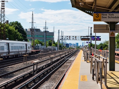 Train Station Platform - A train on the tracks from a train station platform under blue sky and a city skyline in the background 