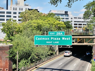 Brooklyn Highway - A road sign on a highway under a bridge with trees and buildings in the background 