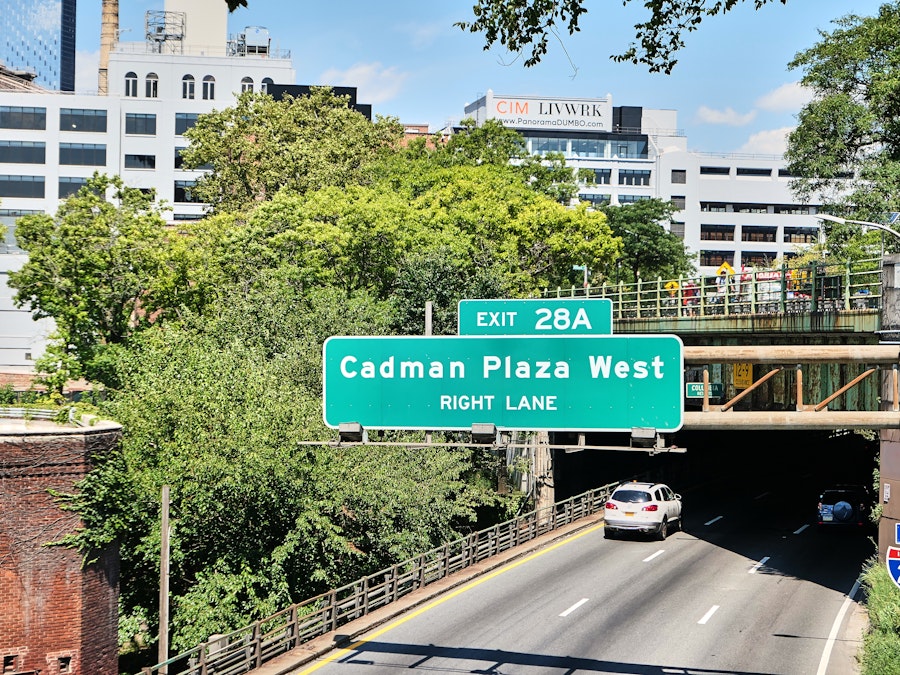 Photo: A road sign on a highway under a bridge with trees and buildings in the background 