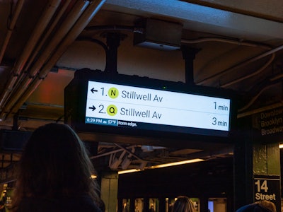 New York City Subway Station Sign - A subway arrivals sign with a couple of people in the background waiting on the platform 