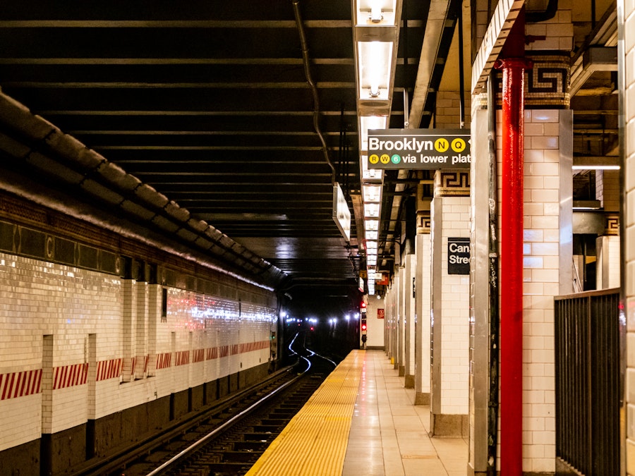 Photo: A subway platform in New York City with tile, overhead lighting, and signs hanging from the ceiling. View of the train tracks.