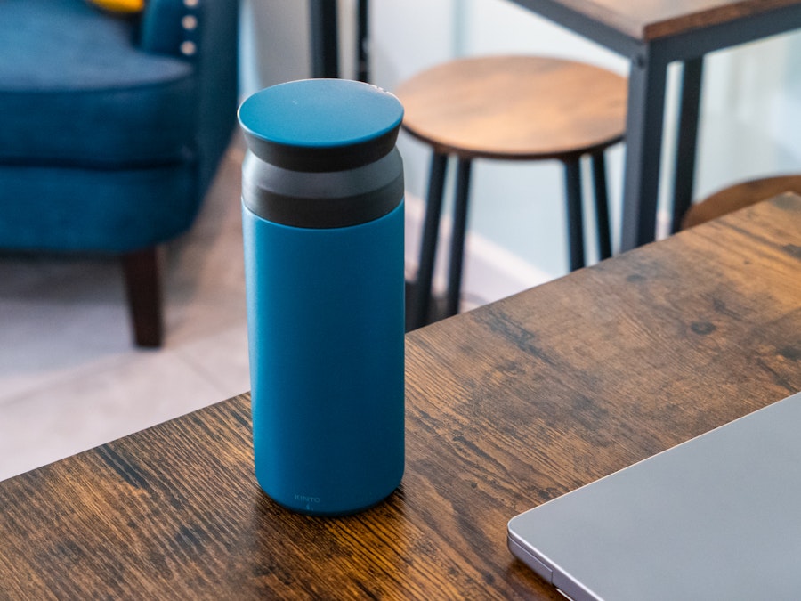 Photo: A blue and black tumbler on a wooden table in a coffee shop with other chairs, couches, and tables blurred in the background. 