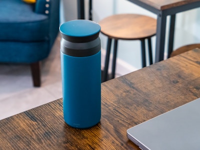 Tumbler and Laptop on Table at Coffee Shop - A blue and black tumbler on a wooden table in a coffee shop with other chairs, couches, and tables blurred in the background. 