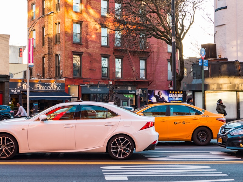 Photo: A white car and yellow taxi on a city street surrounded by buildings and storefronts 