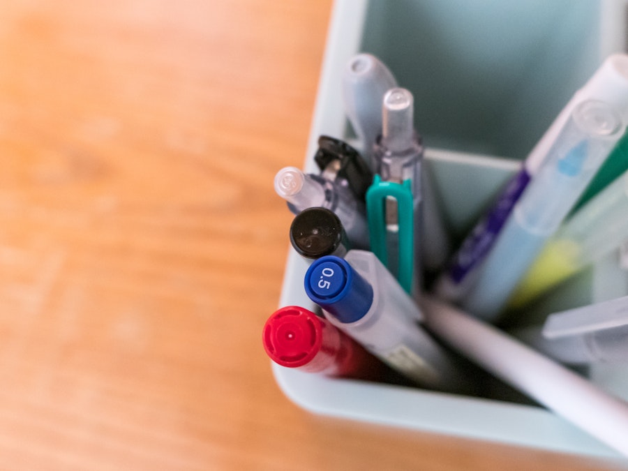 Photo: A group of pens and markers in focus sitting in a container on a wooden desk