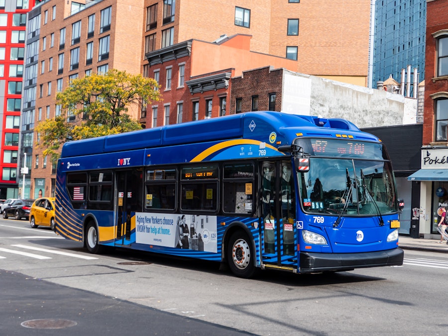 Photo: A blue city bus with a yellow stripe on the side on a city street surrounded by buildings 