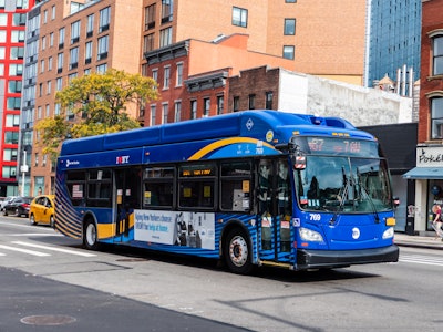Bus on Street in Brooklyn - A blue city bus with a yellow stripe on the side on a city street surrounded by buildings 