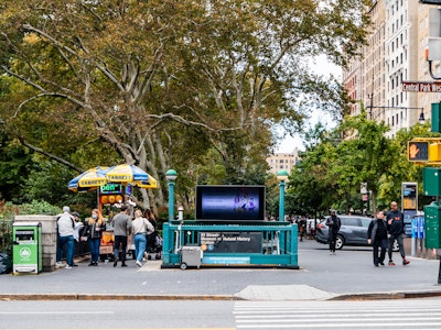 People Outside Central Park and Subway Station - A group of people walking on a city street with a subway entrance in the middle 