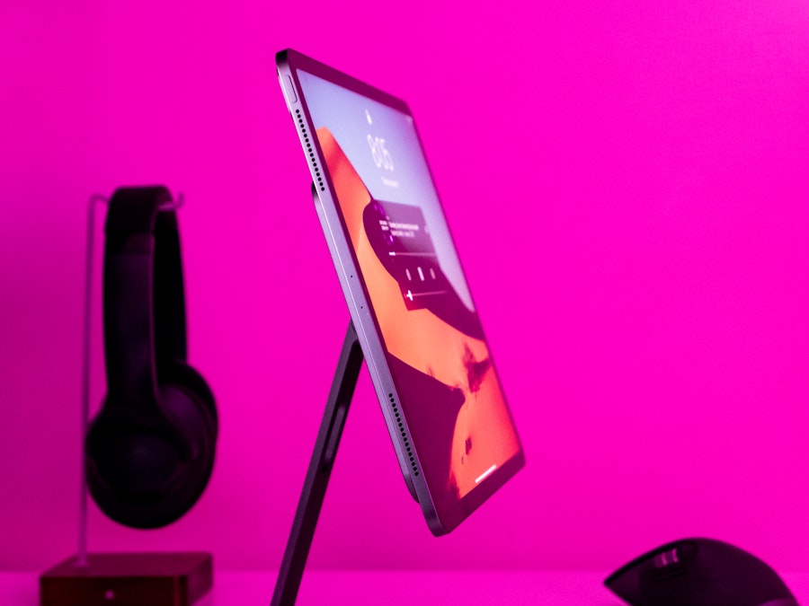 Photo: A tablet on a stand with headphones and a computer mouse in the background under pink lighting 
