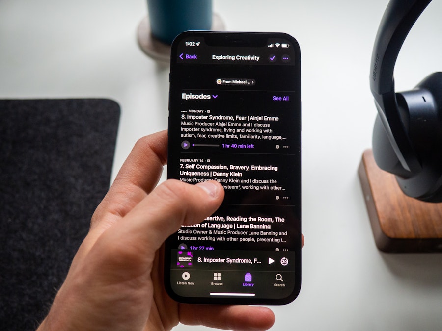 Photo: A person holding a smartphone listening to a podcast over a white desk with headphones in the background 