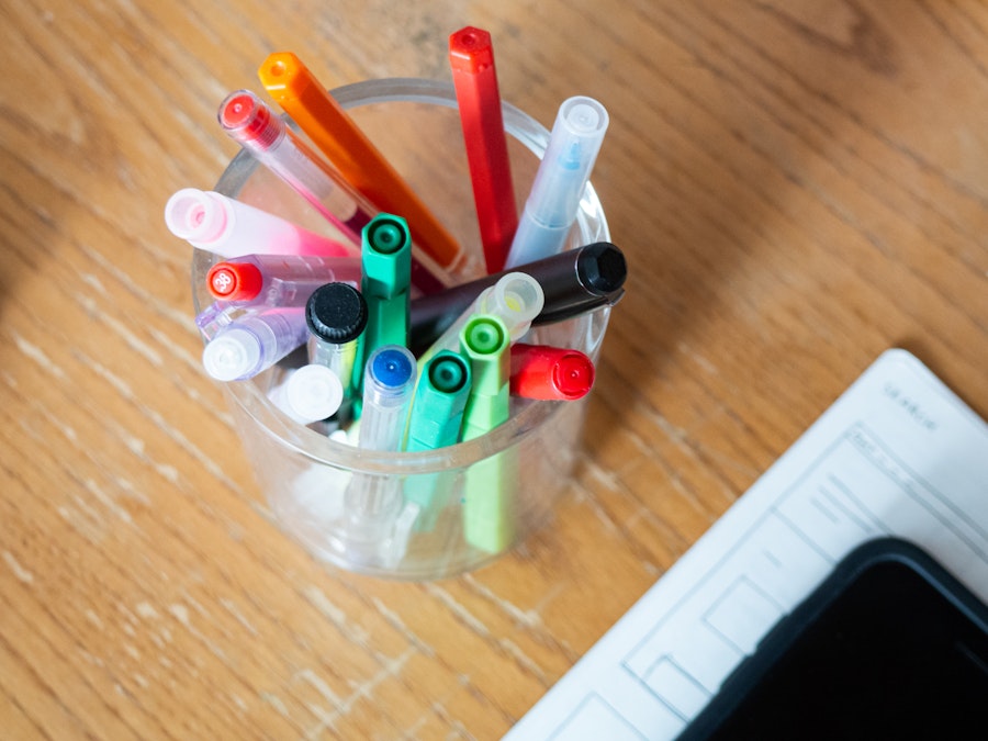 Photo: A cup full of pens, pencils, and markers next to a blurred sketchbook on a wooden desk 