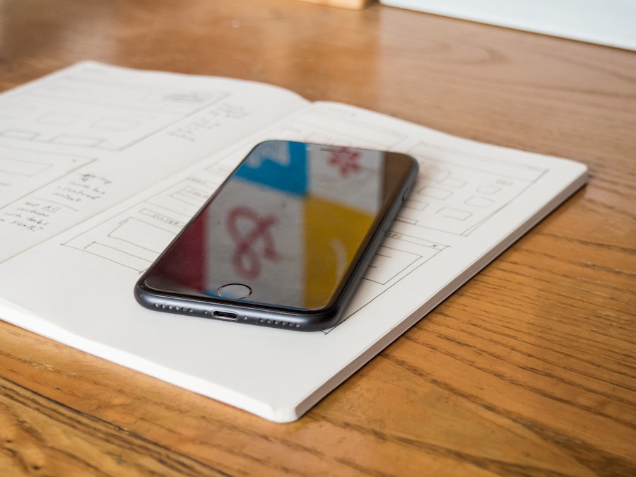 Photo: A smartphone on top of an open sketchbook, on a wooden desk with a poster in the reflection 