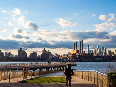 Person Looking Into the Sunset and Manhattan Buildings - People standing and  walking on a boardwalk overlooking a body of water and a city skyline 