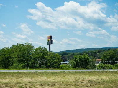 Landscape with Trees and Mountains - A field of grass with a sign on a pole and a road in the background