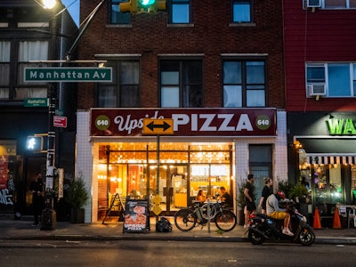 People Outside Brooklyn Pizzeria - A pizzeria with a sign, surrounded by other buildings, and people on the sidewalk